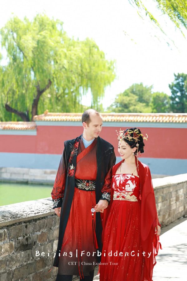 hanfu photoshoot, the forbidden city, beijing, couple, chinatravel, Couple in traditional Chinese wedding attire walking hand in hand along a stone wall beside a red wall and green trees.