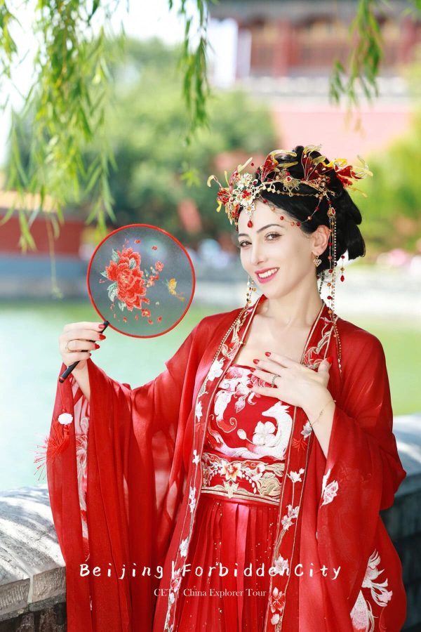 Hanfu photoshoot, the forbidden city, beijing, solo, chinatravel, Woman in a red traditional Chinese dress and ornate headpiece holds a round embroidered fan with a red flower outdoors.