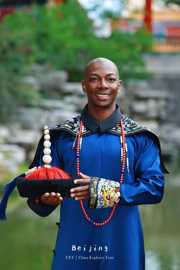 hanfu photoshoot, the forbidden city, beijing, qing dynasty, male, solo, chinatravel, Man in a royal blue traditional Chinese outfit, holding a black fur hat with red tassels, standing outdoors with greenery behind him.