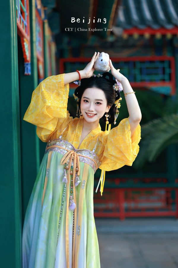 Hanfu photoshoot, the forbidden city, beijing, chinatravel, Smiling woman in a yellow traditional dress posing with hands above her head, holding a white fluffy accessory, in a temple courtyard.