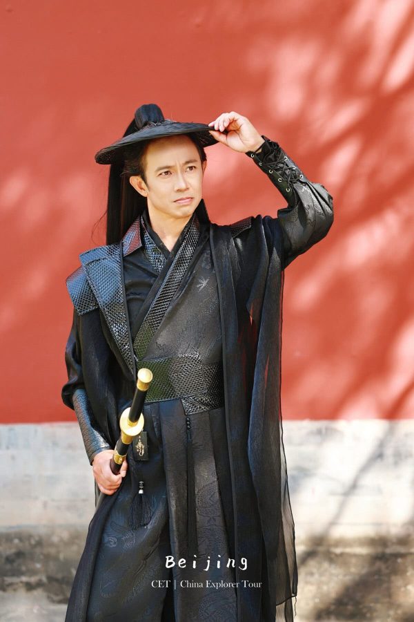 Hanfu photoshoot, the forbidden city, beijing, chinatravel, Man in a black traditional Chinese outfit with a sword, tipping a black hat against a red wall (Beijing travel promo). See bottom text: Beijing CET | China Explorer Tour.