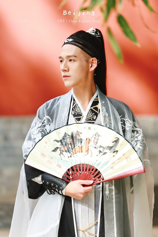 Hanfu Photoshoot, Beijing, zhongshan park, male, chinatravel, Person in traditional Chinese hanfu dress holding a decorative fan, standing against a red background with greenery above