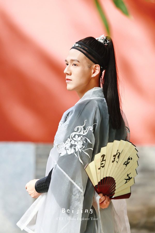 Hanfu Photoshoot, Beijing, zhongshan park, male, chinatravel, Person in traditional Chinese hanfu with embroidered sleeves, holding a folding fan, shown in profile against a peach-colored background.