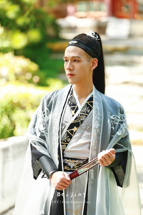 Hanfu Photoshoot, Beijing, zhongshan park, male, chinatravel, Young man wearing ornate traditional Chinese hanfu with a black headband, holding a folding fan outdoors in a garden setting.