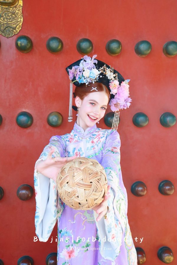 hanfu photoshoot, the forbidden city, beijing, qing dynasty, female, solo, chinatravel, Young woman in a pastel floral hanfu with an ornate flower headdress, extending a woven ball toward the camera against a red gate backdrop.