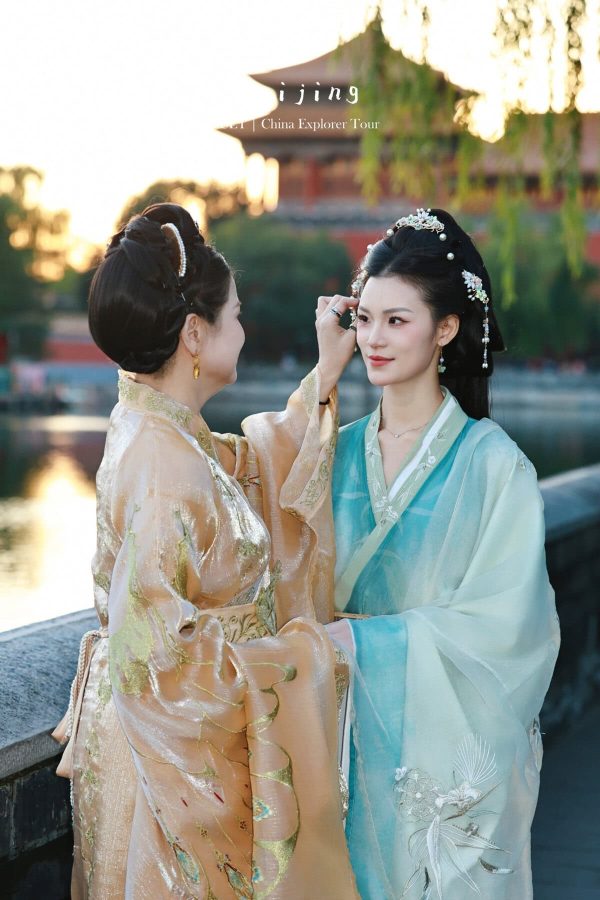 Hanfu photoshoot, Beijing, forbidden city exterior, mother and daughter, chinatravel, Two women in ornate hanfu by a riverside; one gently applies makeup to the other's eye while warm sunset light glows behind them