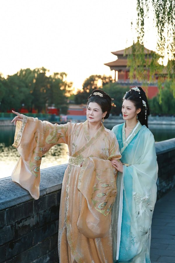 Hanfu photoshoot, Beijing, forbidden city exterior, mother and daughter, chinatravel, Two women in elaborate Hanfu dresses stand at a stone railing by a lake at sunset, with a pagoda in the background and hanging willow branches.