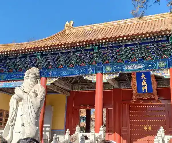 Guozijian,Beijing, chinatrip, Stone statue of a scholar in front of a colorful Chinese temple gate with red columns and ornate blue-green detailing on the roof.