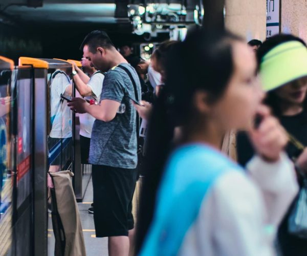 Commuters wait on a subway platform beside a blue train, some looking at phones; a masked woman in the crowd is visible.
