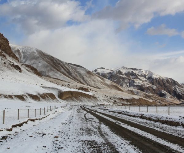 Qiyi Glacier,Jiayuguan,China tour Snow-covered mountain valley with a muddy, tire-tracked road and fence posts running along it, under a blue sky with clouds.
