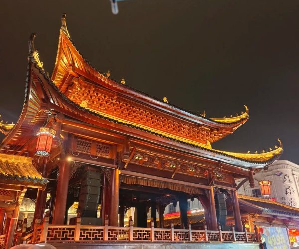 Wanshou Palace,Nanchang,China tour Red-and-gold traditional Chinese temple at night with ornate curved roofs and lanterns; people sit along the front walkway on a wet pavement.