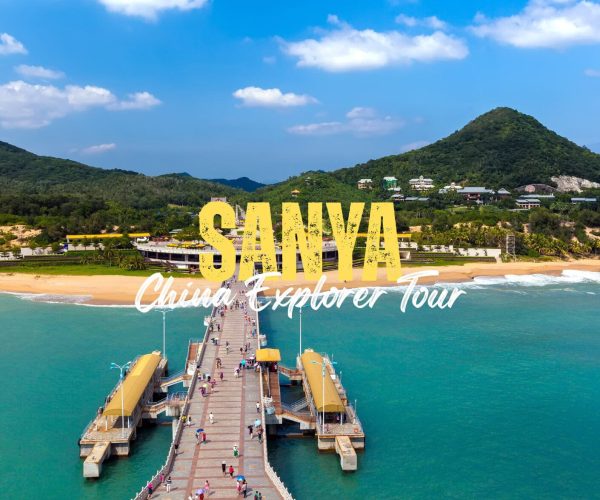Sanya,Hainan,Chinatravel Aerial view of a long pier extending to a sunny Sanya beach with turquoise water and green hills; large yellow 'SANYA' and white 'China Explorer Tour' text overlay.