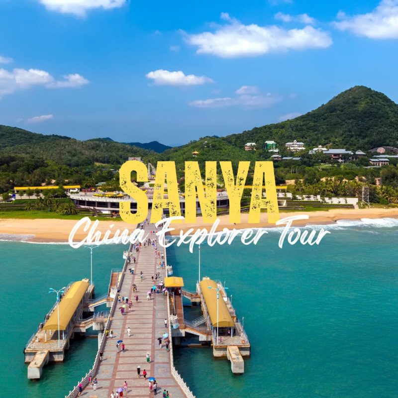 Sanya,Hainan,Chinatravel Aerial view of a long pier extending to a sunny Sanya beach with turquoise water and green hills; large yellow 'SANYA' and white 'China Explorer Tour' text overlay.