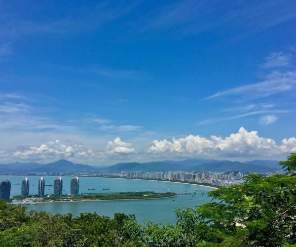 Sanya,Hainan,Chinatravel Four curved glass skyscrapers line a crescent bay, with the city and distant mountains under a clear blue sky, seen from greenery in the foreground.