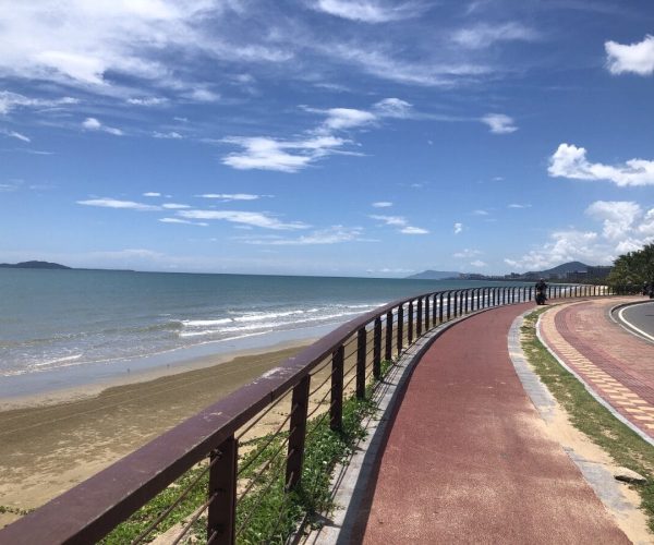 Sanya Bay Coastal Promenade,Sanya,Chinatravel Coastal boardwalk with a red path beside a railing, calm sea, and a blue sky with scattered clouds.