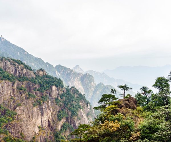 Shangrao Weather,Shangrao,Chinatrip Rocky mountain cliffs covered with trees, fading into a hazy blue horizon under a pale sky.
