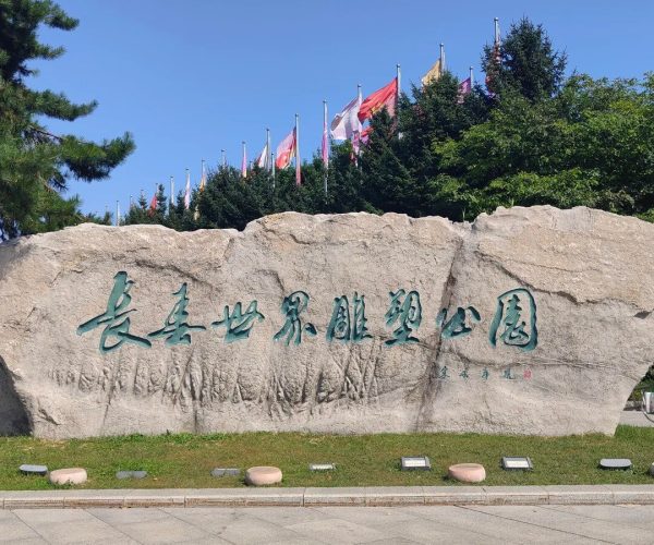 Changchun World Sculpture Park,Changchun,China tour Large stone with turquoise Chinese characters serving as a park sign, with flagpoles and green trees behind under a clear blue sky.