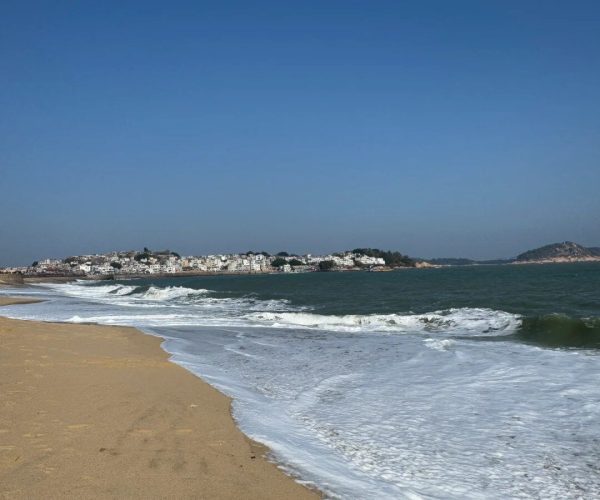 Dongshan Island,Zhangzhou,Chinatravel Wide view of a sandy beach with foamy waves along the shore and a distant town on the horizon under a clear blue sky?