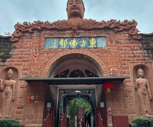 Oriental Buddha Park,Leshan,Chinatravel Temple entrance with a large Buddha head statue atop a red, intricately carved archway and gold Chinese characters above the doorway, flanked by stone statues; visitors enter through an open gate.