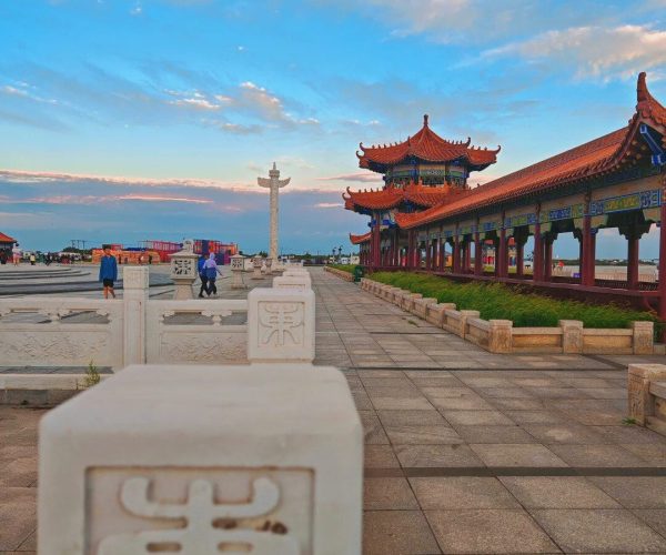East Pole Square & Wusu Town,Jiamusi,Chinatravel White stone railing with a carved Chinese character in the foreground, leading to a red-roofed Chinese temple pavilion along a paved walkway.