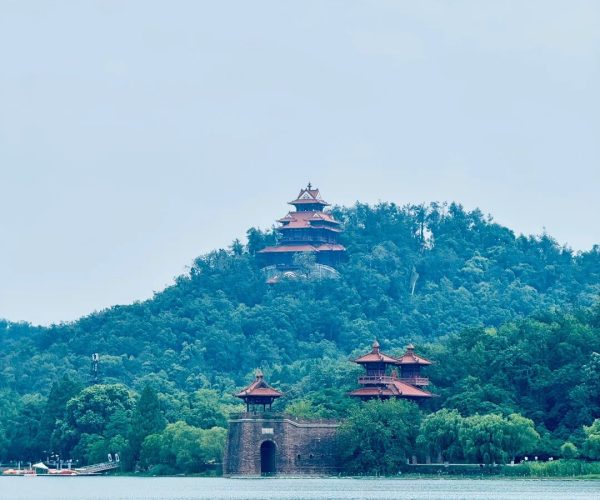 East Lake Scenic Area,Wuhan,Chinatravel Three-tiered red pagoda perched on a forested hill beside a calm lake, with a stone gate at the water's edge.