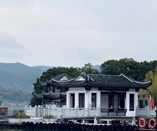 Dongqian Lake,Ningbo,Chinatravel White pavilion with dark curved roofs on a calm lake, connected by a white railing and floating tires along the edge, mountains in the distance