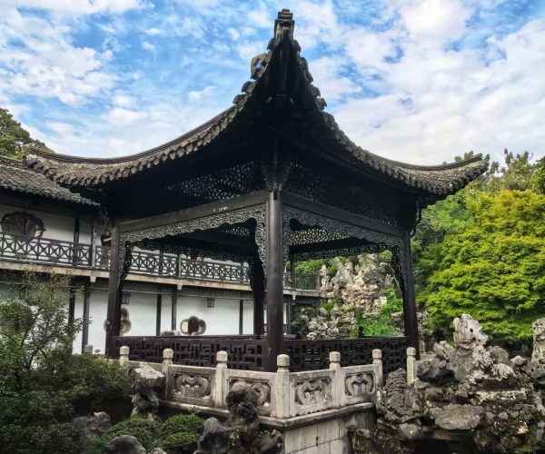 Geyuan Garden,Yangzhou,China tour Black ornate Chinese pavilion on a stone platform over water, surrounded by rocks and greenery under a blue sky with clouds