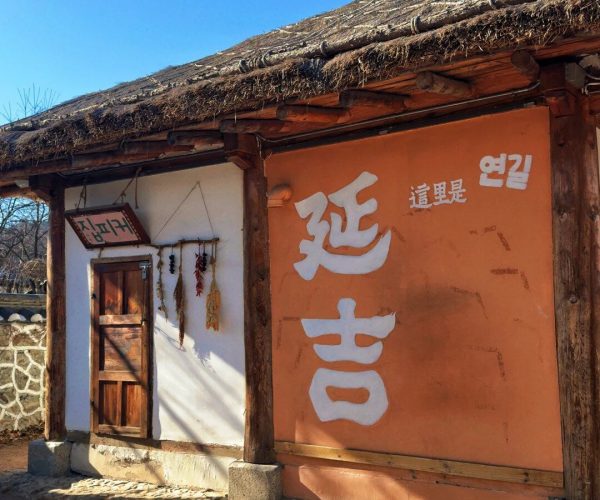 Korean Folk Culture Park,Jilin Province,China tour Traditional Korean countryside hut with a thatched roof, white door, and a large orange wall featuring white Korean characters; dried herbs hang by the doorway.