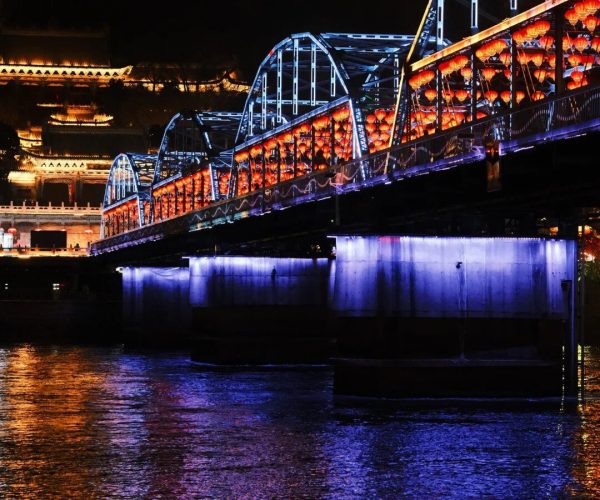 Night view of a blue lit bridge adorned with red lanterns over a dark river, with illuminated traditional buildings in the background.