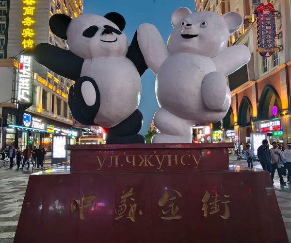 Sino-Soviet Golden Street,Manzhouli,China tour Two large cartoon panda and white bear statues high-fiving atop a red pedestal in a neon-lit street plaza with Chinese signage behind them