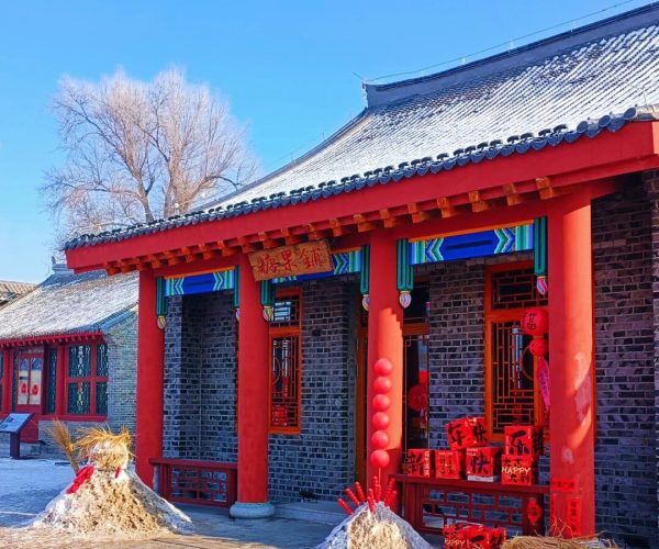 Wulajie Manchu Village,Jilin,Chinatrip Red-painted traditional Chinese building doorway with red columns and festive decorations; red lanterns, banners, and gift boxes outside against a blue sky. A snow pile with straw decorations sits nearby.