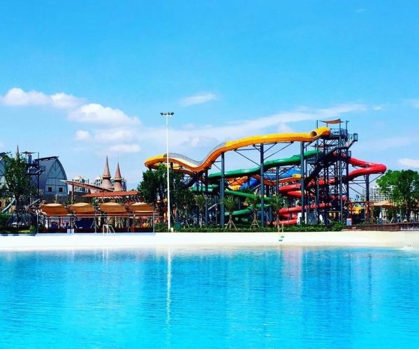 Leshan Happy Water World,Leshan,Chinatravel Colorful winding water slides at a sunny outdoor water park beside a large blue pool in front of palm trees and buildings.