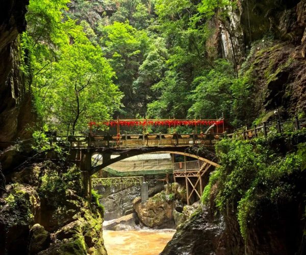 Jiuxiang Scenic Area ,Kunming,Chinatrip View of a green canyon with a wooden bridge adorned with red lanterns spanning a brown rushing river.