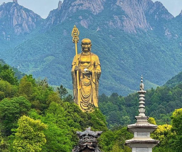 Jiuhua Mountain,Chizhou,China tour Giant golden Buddha statue with a staff in a forested mountain park, beside a stone pagoda and dragon sculpture.