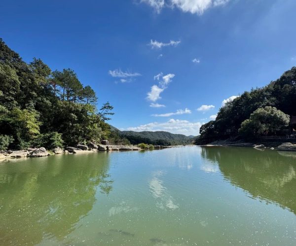Jiuli Lake,Putian,Chinatravel Calm river flanked by green trees and rocky banks under a bright blue sky with fluffy clouds; reflections ripple on the water surface.