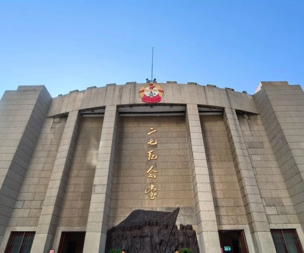 Erqi Memorial Tower,Zhengzhou,Chinatravel Large gray stone building with tall arches and golden vertical Chinese characters on the façade, a crest atop the center, and a bas-relief sculpture near the entrance against a clear blue sky