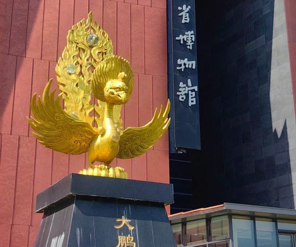 Yunnan Provincial Museum,Kunming,Chinatravel Golden phoenix statue with outstretched wings on a dark pedestal outside a red-brick building.