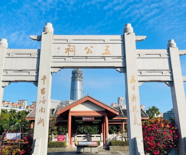 Temple of the Five Lords,Haikou,Chinatravel Stone archway with Chinese inscriptions frames a traditional pavilion; a tall modern skyscraper rises in the background under a blue sky.