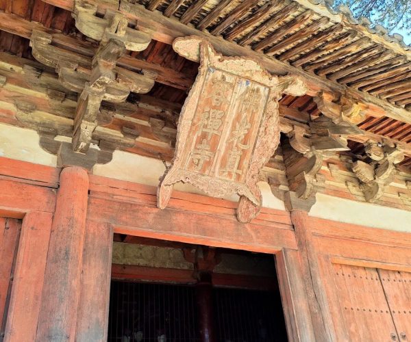 Foguang Temple,Xinzhou,Chinatravel Old wooden temple entrance with a carved, weathered signboard hanging above the doorway; red columns and exposed beams under a tile roof.