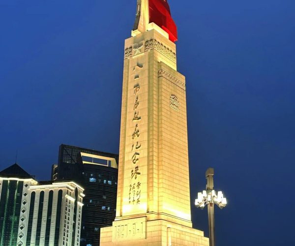 Bayi Square,Nanchang,China tour Tall illuminated obelisk monument at night with a red flag on top and Chinese inscriptions, surrounded by modern buildings.