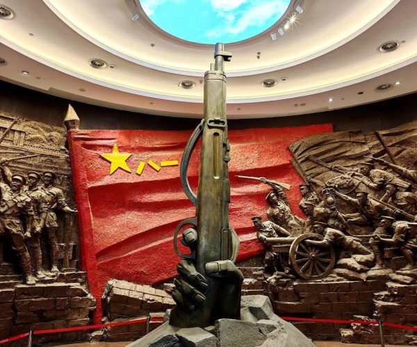 August 1st Nanchang Uprising Museum,Nanchang,China tour A towering rifle sculpture planted in jagged rocks, set against a red war relief wall with soldiers and a cannon wheel in a museum display.
