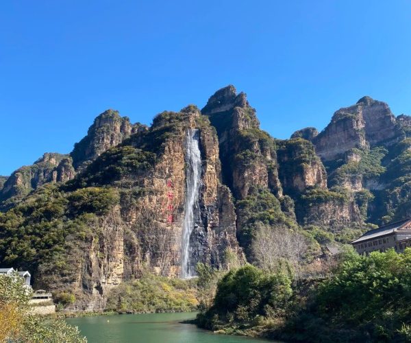 Xinglong Mountain National Nature Reserve,Lanzhou,China tour Waterfall on a rocky cliff cascading into a green river, with blue sky and surrounding mountains nearby buildings on the right.