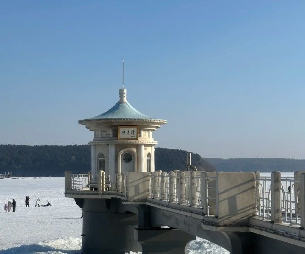 Jingyuetan National Forest Park,Changchun,China tour Pavilion with a teal pagoda roof at the end of a walkway extending over a frozen lake, under a clear blue sky.