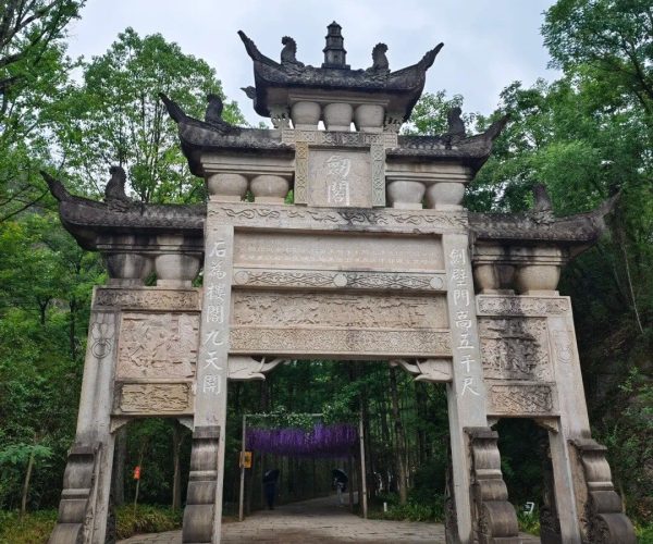 Cantonment Ancient Town,Mianyang,Chinatravel Stone ornate Chinese paifang gate with carved panels and decorative upturned roofs, set in a green forest with a paved path beyond.