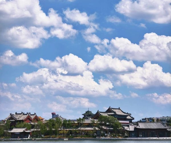 Baogong Lake & Park,Kaifeng,Chinatravel Waterfront view of traditional Chinese buildings along a lake, with trees and a blue sky filled with white clouds behind them.