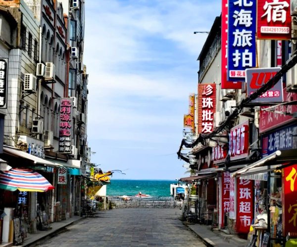 Beihai Old Street,Beihai,Chinatravel Narrow stone-paved street in a seaside town, flanked by buildings with colorful Chinese signboards, leading to a blue sea under a clear sky.