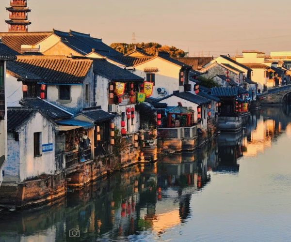 Qiandeng Ancient Town,Kunshan,China tour Sunlit canal town with white-walled houses, dark tiled roofs, red lanterns, and a pagoda reflected in calm water.