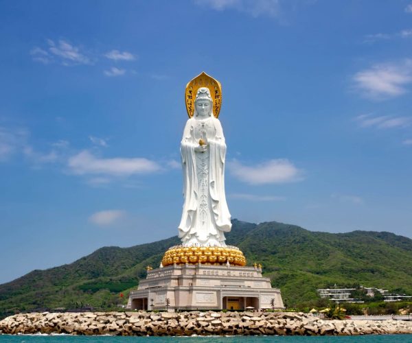 Nanshan Temple & Giant Guanyin Statue,Sanya,Chinatravel Large white Guanyin statue standing on a golden lotus pedestal by the sea, with green hills in the background.