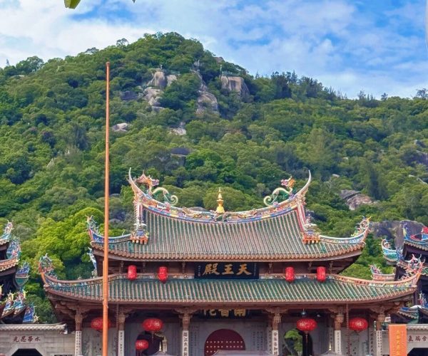 Nanputuo Temple,Xiamen,Chinatravel Colorful Chinese temple with ornate curved roofs and red lanterns at the base of a green hillside under a blue sky, crowds of people gathered outside.