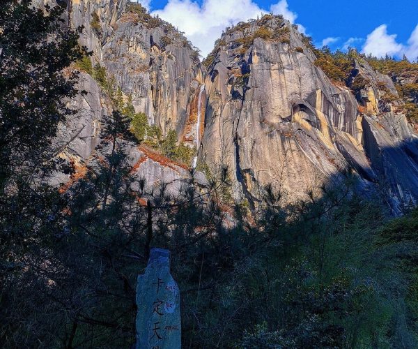 Kading Creek,Nyingchi,China tour Tall rock cliffs in a deep canyon under a bright blue sky; foreground trees and a stone monument with Chinese inscriptions.